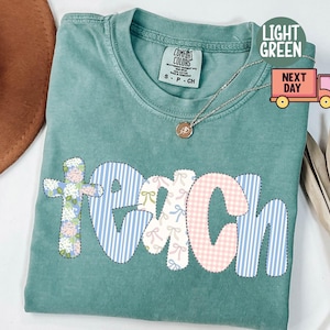 May include: Light green Comfort Colors t-shirt with the word "TEACH" in a patchwork design. The letters are decorated with floral, striped, and gingham patterns. A gold necklace with a pendant is visible.