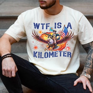 May include: Cream-colored t-shirt with a graphic featuring an eagle carrying a soldier with a rifle and American flag. The text "WTF IS A KILOMETER" is printed in blue. The shirt is worn by a person.
