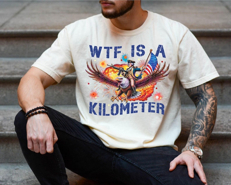 May include: Cream-colored t-shirt with a graphic featuring an eagle carrying a soldier with a rifle and American flag. The text "WTF IS A KILOMETER" is printed in blue. The shirt is worn by a person.
