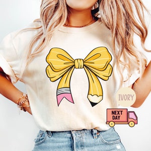 Può includere: Una maglietta bianca con un design a fiocco di matita giallo. Il fiocco è legato con una gomma da cancellare rosa. La maglietta è etichettata "Avorio" e "Il giorno dopo".