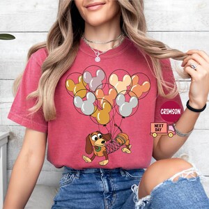 May include: A red t-shirt with a cartoon dog holding balloons shaped like Mickey Mouse ears. The balloons are in shades of brown, orange, yellow, and white. The dog is wearing a blue collar and has a spring attached to its tail.