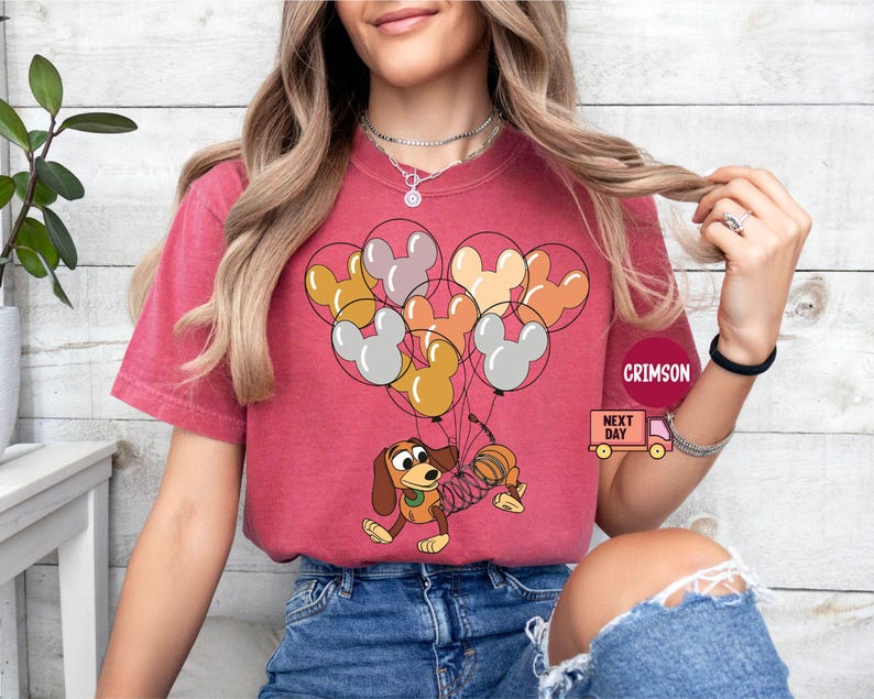 May include: A red t-shirt with a cartoon dog holding balloons shaped like Mickey Mouse ears. The balloons are in shades of brown, orange, yellow, and white. The dog is wearing a blue collar and has a spring attached to its tail.