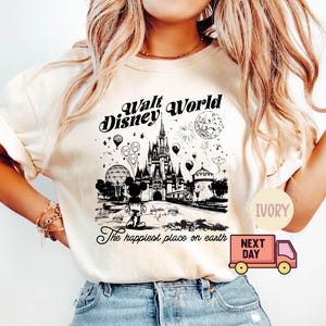 May include: Ivory-colored t-shirt featuring a black and white graphic of Walt Disney World. The design includes Cinderella's castle, Mickey Mouse, balloons, and the text "The happiest place on earth."