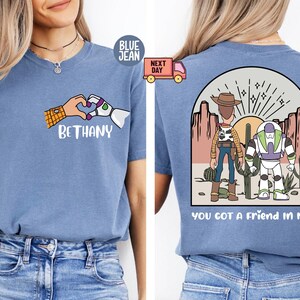 Pu&ograve; includere: Una t-shirt blu con una grafica frontale di una mano umana e una mano robotica che formano un cuore sopra il nome BETHANY. La grafica sul retro mostra due personaggi dei cartoni animati in un deserto con il testo YOU GOT A FRIEND IN ME.