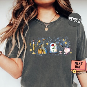 May include: A dark gray t-shirt with a graphic print of a castle, a rose in a glass dome, a candlestick, a book, and a teapot. The graphic is in a cartoon style and has a blue and yellow color scheme. The shirt has a distressed look and is made of a soft, comfortable material.