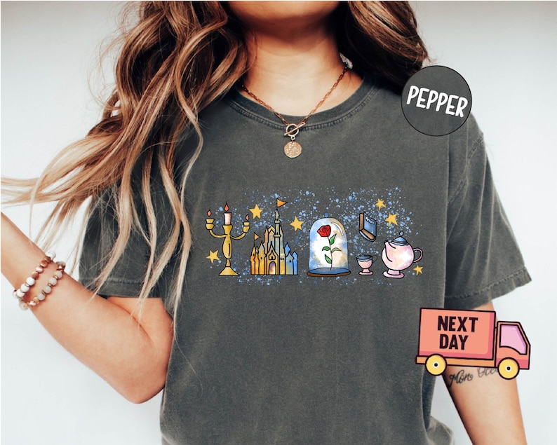 May include: A dark gray t-shirt with a graphic print of a castle, a rose in a glass dome, a candlestick, a book, and a teapot. The graphic is in a cartoon style and has a blue and yellow color scheme. The shirt has a distressed look and is made of a soft, comfortable material.