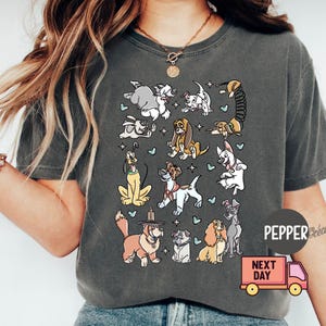 May include: Dark gray t-shirt featuring a colorful cartoon illustration of various dog breeds. The design includes dogs of different sizes and colors, with small blue heart accents. The shirt has a relaxed fit.