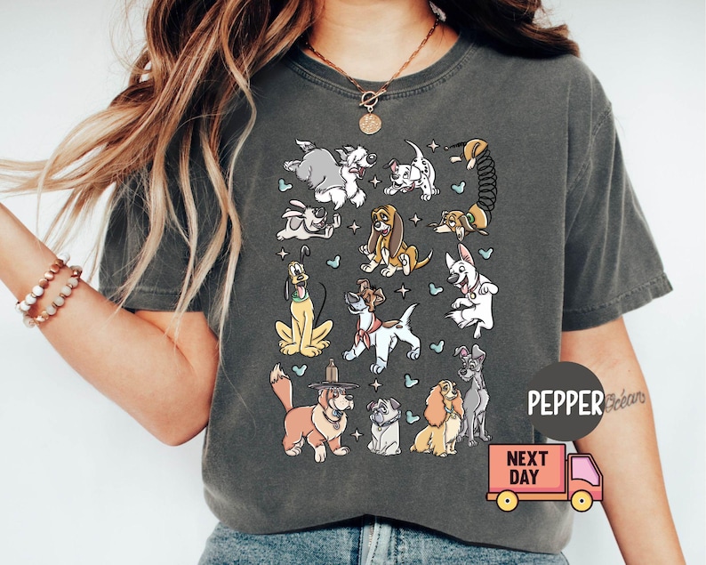 May include: Dark gray t-shirt featuring a colorful cartoon illustration of various dog breeds. The design includes dogs of different sizes and colors, with small blue heart accents. The shirt has a relaxed fit.