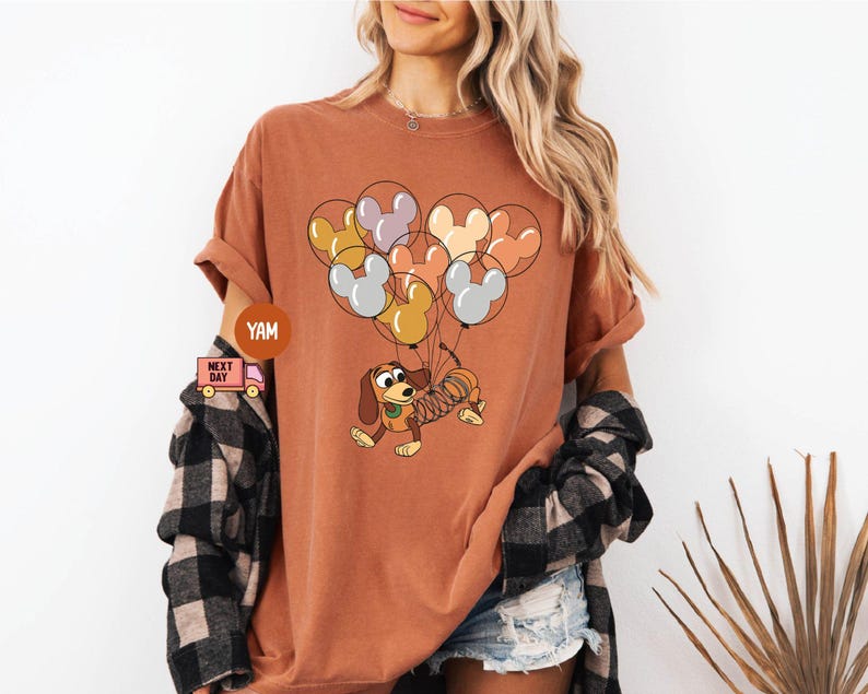 May include: A rust-colored t-shirt with a cartoon image of a dachshund dog holding onto a bunch of balloons. The balloons are shaped like Mickey Mouse ears and are in shades of gray, brown, and yellow.