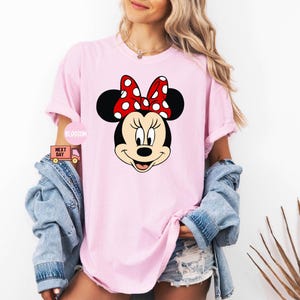 Może przedstawiać: R&oacute;żowy T-shirt z kresk&oacute;wkowym wizerunkiem Minnie Mouse. Minnie Mouse ma na sobie czerwoną kokardkę w białe kropki i ma czarny nos i oczy.