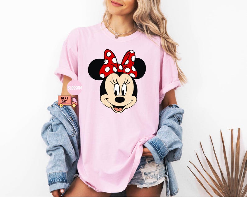 Może przedstawiać: R&oacute;żowy T-shirt z kresk&oacute;wkowym wizerunkiem Minnie Mouse. Minnie Mouse ma na sobie czerwoną kokardkę w białe kropki i ma czarny nos i oczy.