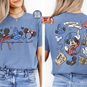 Puede incluir: Camiseta vaquera azul con un diseño gráfico frontal que presenta orejas de Mickey Mouse, un sombrero de mago y un lazo de Minnie Mouse. El texto "Some imagination, huh?" también está presente. La parte trasera de la camiseta tiene un diseño de Mickey Mouse.