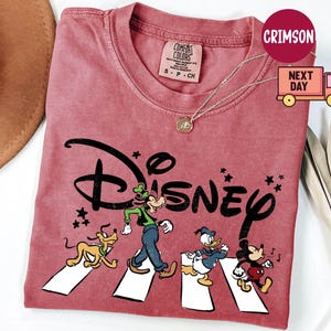K&ouml;nnte beinhalten: Ein staubrosa T-Shirt mit dem Wort "Disney" in schwarzer, stilisierter Schrift. Das Shirt zeigt die Zeichentrickfiguren Goofy, Pluto, Donald Duck und Mickey Mouse, die einen Zebrastreifen &uuml;berqueren. Eine goldene Halskette ist sichtbar.