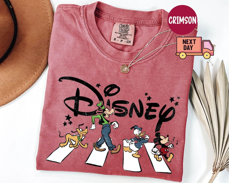 K&ouml;nnte beinhalten: Ein staubrosa T-Shirt mit dem Wort "Disney" in schwarzer, stilisierter Schrift. Das Shirt zeigt die Zeichentrickfiguren Goofy, Pluto, Donald Duck und Mickey Mouse, die einen Zebrastreifen &uuml;berqueren. Eine goldene Halskette ist sichtbar.