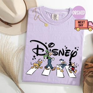K&ouml;nnte beinhalten: Ein helllila T-Shirt mit dem Disney-Logo und Zeichentrickfiguren, die eine Stra&szlig;e &uuml;berqueren. Das T-Shirt ist mit einem Hut, Sandalen und einer Halskette versehen. Das T-Shirt hat den Text "ORCHID" und eine Grafik eines Lieferwagens.