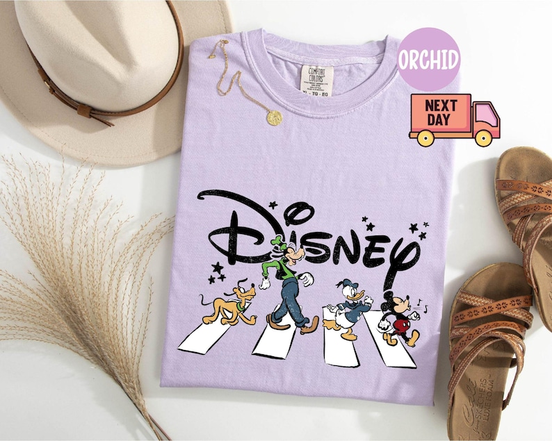 K&ouml;nnte beinhalten: Ein helllila T-Shirt mit dem Disney-Logo und Zeichentrickfiguren, die eine Stra&szlig;e &uuml;berqueren. Das T-Shirt ist mit einem Hut, Sandalen und einer Halskette versehen. Das T-Shirt hat den Text "ORCHID" und eine Grafik eines Lieferwagens.