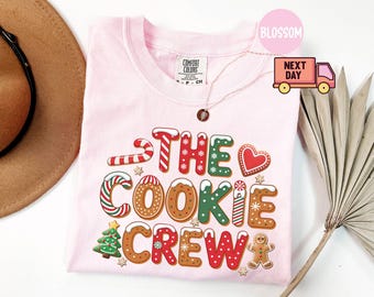 May include: A light pink Comfort Colors t-shirt with the text "THE COOKIE CREW" in a colorful, gingerbread-style font. The design includes gingerbread men, candy canes, and a Christmas tree. A gold necklace is draped over the shirt.