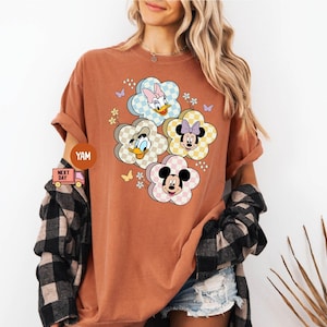 May include: A rust-colored t-shirt featuring a floral design with Disney characters. The design includes Mickey Mouse, Minnie Mouse, Donald Duck, and Daisy Duck, each within a flower-shaped frame with a checkered pattern.