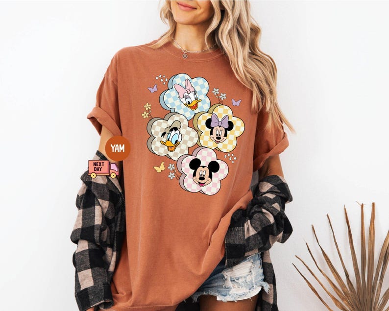 May include: A rust-colored t-shirt featuring a floral design with Disney characters. The design includes Mickey Mouse, Minnie Mouse, Donald Duck, and Daisy Duck, each within a flower-shaped frame with a checkered pattern.