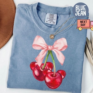 Puede incluir: Una camiseta vaquera azul con un dise&ntilde;o de acuarela de cerezas y un lazo rosa. Las cerezas son rojas con detalles en negro. La camiseta tiene una etiqueta Comfort Colors.