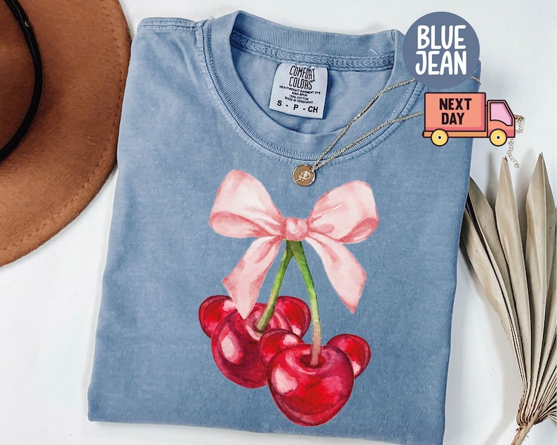 Puede incluir: Una camiseta vaquera azul con un dise&ntilde;o de acuarela de cerezas y un lazo rosa. Las cerezas son rojas con detalles en negro. La camiseta tiene una etiqueta Comfort Colors.
