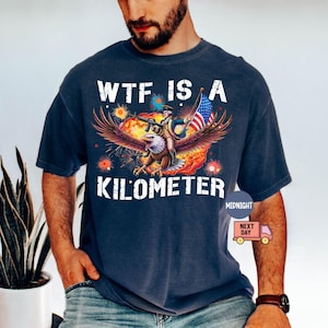 May include: Navy blue t-shirt with a graphic featuring an eagle carrying a person holding a flag. The text "WTF IS A KILOMETER" is printed in white. The design includes red, orange, and yellow accents.