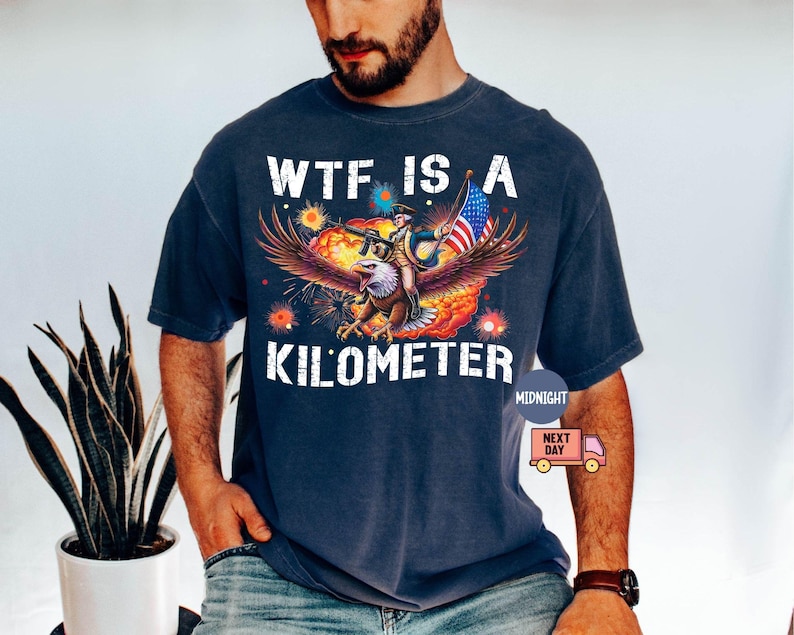 May include: Navy blue t-shirt with a graphic featuring an eagle carrying a person holding a flag. The text "WTF IS A KILOMETER" is printed in white. The design includes red, orange, and yellow accents.