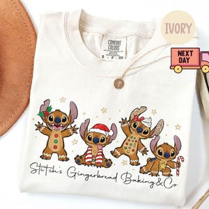 Puede incluir: Camiseta de color marfil con un dise&ntilde;o festivo de cuatro personajes Stitch estilo pan de jengibre. Los personajes est&aacute;n decorados con accesorios navide&ntilde;os y rodeados de estrellas. El texto "Stitch's Gingerbread Baking & Co" est&aacute; impreso debajo.