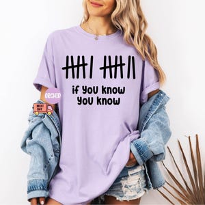 May include: A light purple t-shirt with the text "|||| |||| if you know you know" in black. The shirt is a relaxed fit and has short sleeves. A denim jacket is draped over the shoulders.