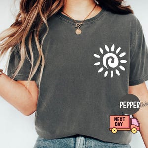 May include: Dark gray t-shirt with a white sun graphic on the front. The sun design features a spiral center and radiating lines. The shirt has a crew neck and short sleeves.