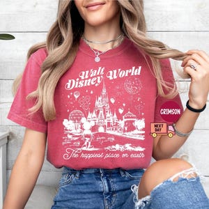 May include: Crimson-colored t-shirt featuring a white graphic of the Walt Disney World castle, Mickey Mouse, and balloons. The text "Walt Disney World" and "The happiest place on earth" are also printed on the shirt.