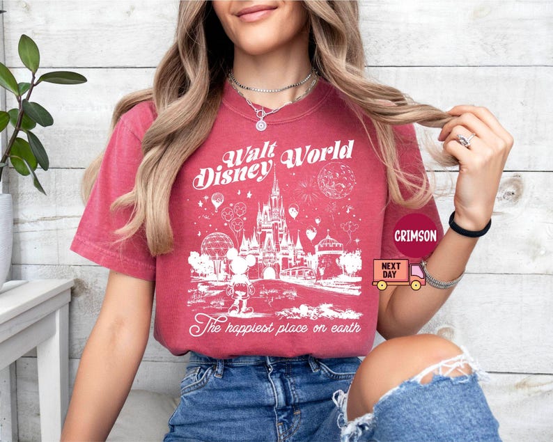 May include: Crimson-colored t-shirt featuring a white graphic of the Walt Disney World castle, Mickey Mouse, and balloons. The text "Walt Disney World" and "The happiest place on earth" are also printed on the shirt.