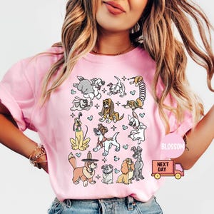 May include: Pink t-shirt featuring a playful illustration of various dog breeds in a repeating pattern. The dogs are depicted in a cartoon style with different colors and poses. The shirt has short sleeves and a crew neck.