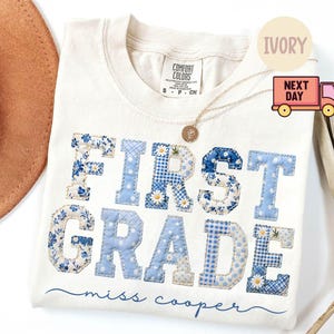 Puede incluir: Una camiseta de manga corta color marfil con el texto FIRST GRADE en letras grandes de estilo bloque, con un patrón de patchwork floral y de cuadros vichy en azul y blanco. Debajo, el nombre miss cooper en letra cursiva azul.