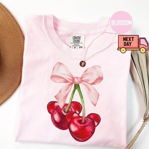 Puede incluir: Una camiseta rosa claro con un dise&ntilde;o de acuarela de cerezas y un lazo rosa. Las cerezas son de color rojo intenso con tallos verdes. La camiseta tiene cuello redondo y mangas cortas. La etiqueta de la marca es visible arriba a la izquierda.