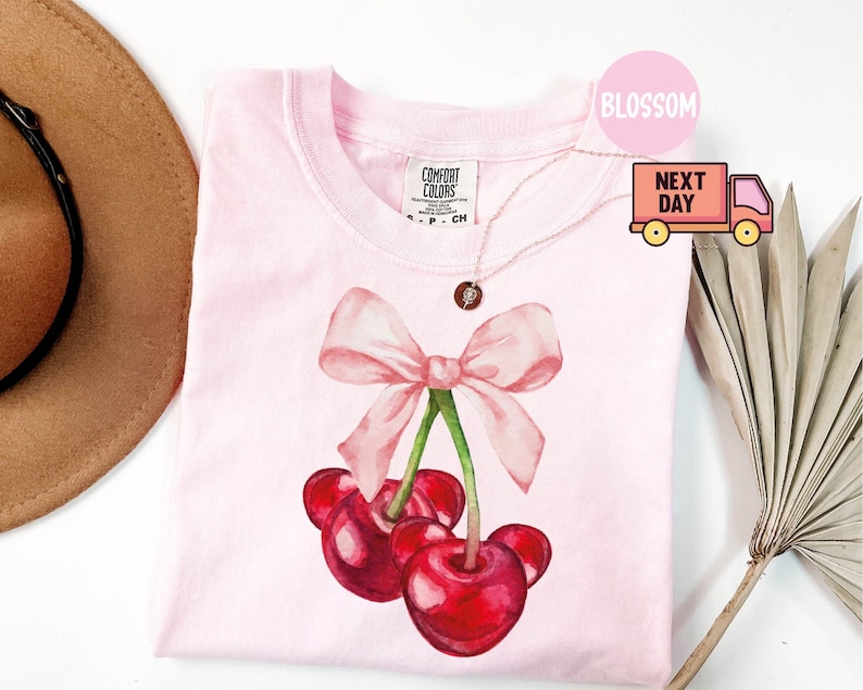 Puede incluir: Una camiseta rosa claro con un dise&ntilde;o de acuarela de cerezas y un lazo rosa. Las cerezas son de color rojo intenso con tallos verdes. La camiseta tiene cuello redondo y mangas cortas. La etiqueta de la marca es visible arriba a la izquierda.