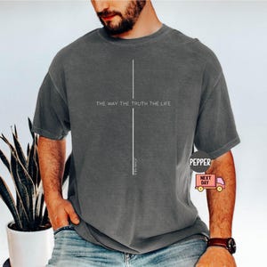 May include: Dark gray t-shirt with the text "THE WAY THE TRUTH THE LIFE" and a vertical line design. The shirt is worn by a person and is paired with blue jeans. The shirt also has the text "JOHN 14:6".