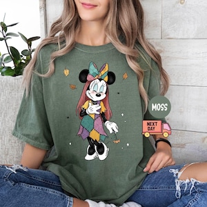 May include: Olive green t-shirt featuring a Minnie Mouse graphic in a patchwork design. The cartoon character has a colorful bow and dress, with autumn leaves accents. The shirt is worn by a person wearing ripped blue jeans.