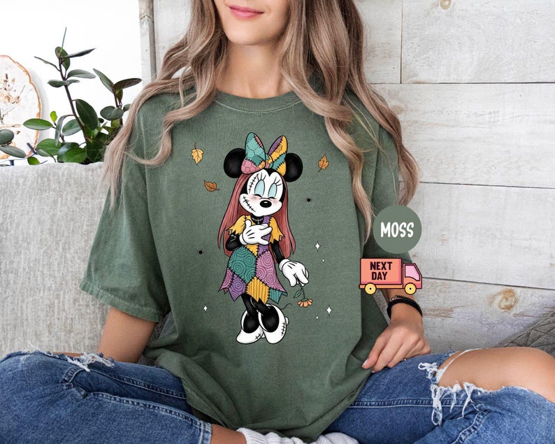 May include: Olive green t-shirt featuring a Minnie Mouse graphic in a patchwork design. The cartoon character has a colorful bow and dress, with autumn leaves accents. The shirt is worn by a person wearing ripped blue jeans.