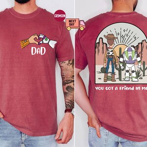 Pu&ograve; includere: Una t-shirt a maniche corte color cremisi con una grafica frontale di due mani che formano un cuore con la parola DAD sotto. La grafica sul retro mostra due personaggi dei cartoni animati in un paesaggio desertico con il testo YOU GOT A FRIEND IN ME.