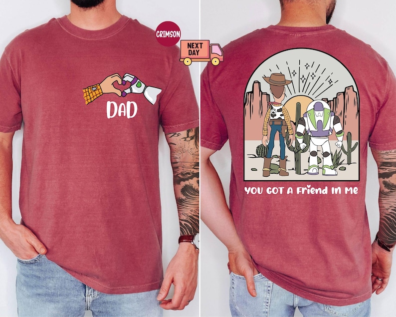 Pu&ograve; includere: Una t-shirt a maniche corte color cremisi con una grafica frontale di due mani che formano un cuore con la parola DAD sotto. La grafica sul retro mostra due personaggi dei cartoni animati in un paesaggio desertico con il testo YOU GOT A FRIEND IN ME.