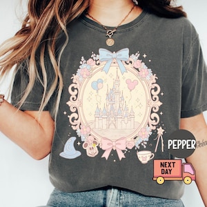 K&ouml;nnte beinhalten: Ein graues T-Shirt mit einem pastellrosa und blauen Motiv von Schloss Aschenputtel, Mickey-Maus-Ohren und einer Schleife. Der Text "Next Day" und "Pepper" befindet sich ebenfalls auf dem Shirt.