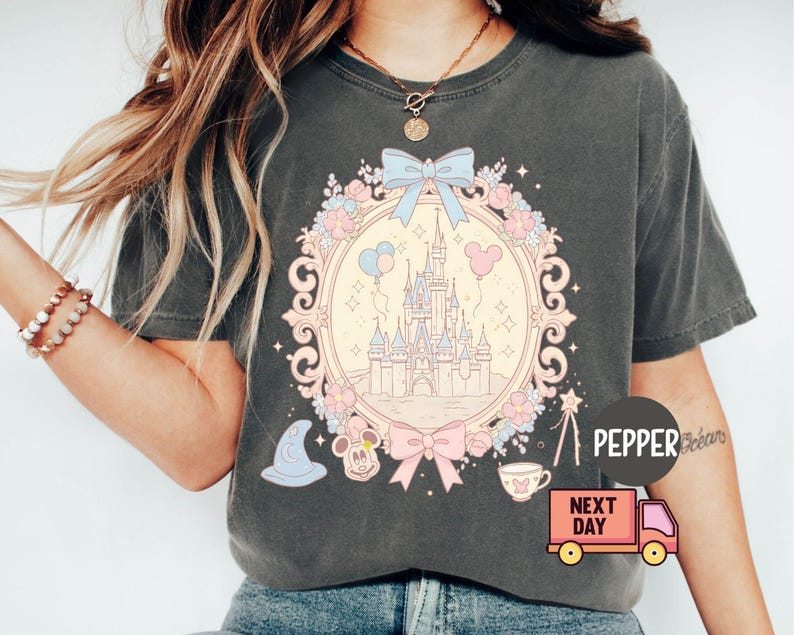 K&ouml;nnte beinhalten: Ein graues T-Shirt mit einem pastellrosa und blauen Motiv von Schloss Aschenputtel, Mickey-Maus-Ohren und einer Schleife. Der Text "Next Day" und "Pepper" befindet sich ebenfalls auf dem Shirt.