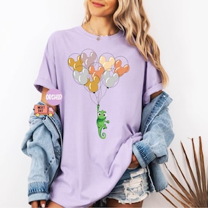 Może przedstawiać: Fioletowy T-shirt z zielonym kameleonem trzymającym wiązkę kolorowych balon&oacute;w w kształcie uszu Myszki Miki. Balony są pomarańczowe, ż&oacute;łte, r&oacute;żowe i szare.