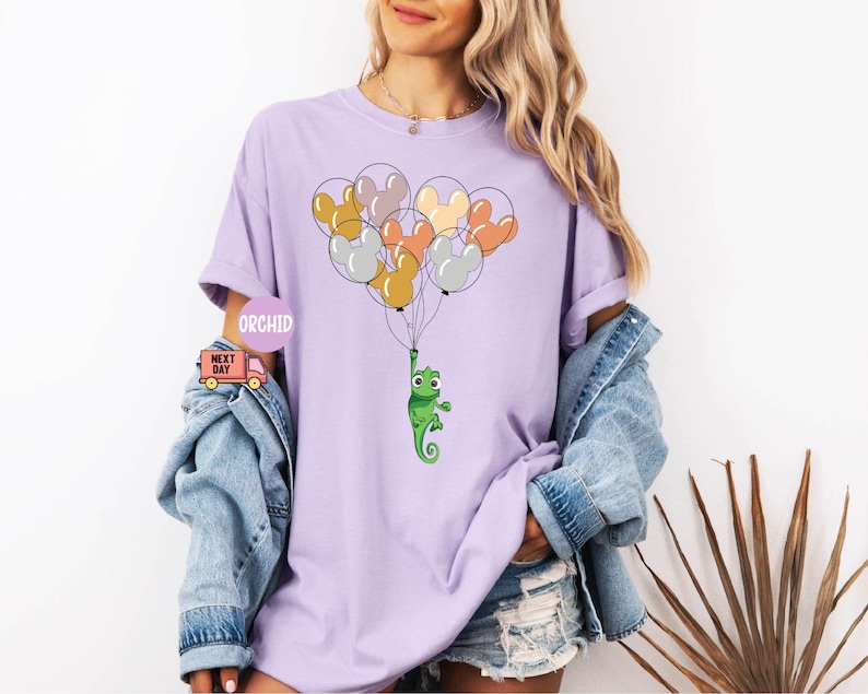 Może przedstawiać: Fioletowy T-shirt z zielonym kameleonem trzymającym wiązkę kolorowych balon&oacute;w w kształcie uszu Myszki Miki. Balony są pomarańczowe, ż&oacute;łte, r&oacute;żowe i szare.