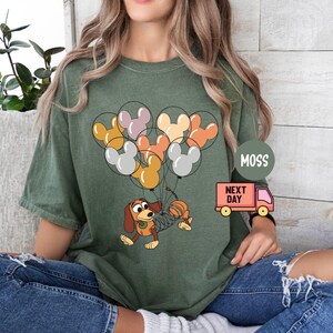 May include: A green t-shirt with a cartoon dog holding colorful balloons shaped like Mickey Mouse heads. The balloons are orange, brown, pink, and white. The text "Next Day" is on a red truck.