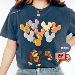 May include: A navy blue t-shirt with a graphic of a cartoon dog holding balloons shaped like Mickey Mouse heads. The balloons are in various colors, including yellow, orange, pink, and gray. The text "Next Day" is printed on a red truck in the bottom right corner.