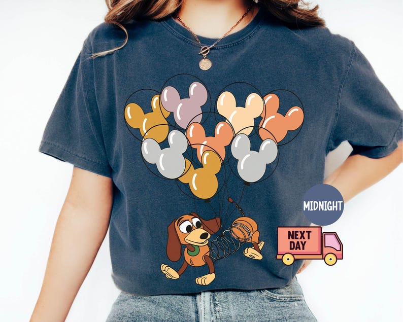May include: A navy blue t-shirt with a graphic of a cartoon dog holding balloons shaped like Mickey Mouse heads. The balloons are in various colors, including yellow, orange, pink, and gray. The text "Next Day" is printed on a red truck in the bottom right corner.