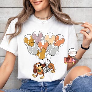 May include: White t-shirt with a cartoon image of a dog holding onto a bunch of colorful Mickey Mouse balloons. The text "Next Day" is printed on a small truck in the bottom right corner of the shirt.