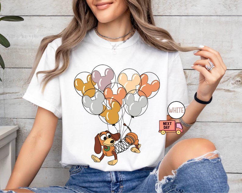 May include: White t-shirt with a cartoon image of a dog holding onto a bunch of colorful Mickey Mouse balloons. The text "Next Day" is printed on a small truck in the bottom right corner of the shirt.